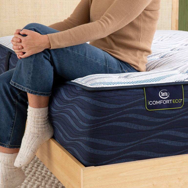 Person sitting on bed to showcase firmness level of Serta iComforteco plush mattress||feel: plush||level: enhanced