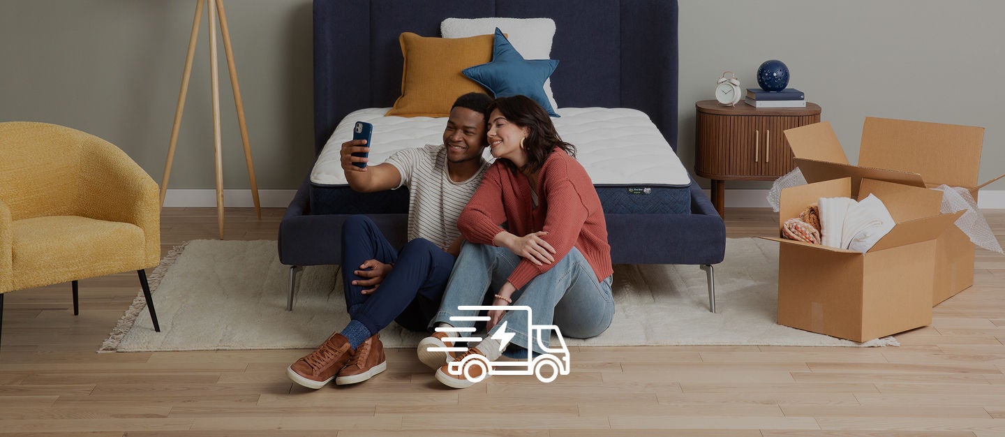 Man and woman sitting on the flow in front of their Serta mattress in a blue bedframe taking a self portrait with open boxes on the floor to the right of them.