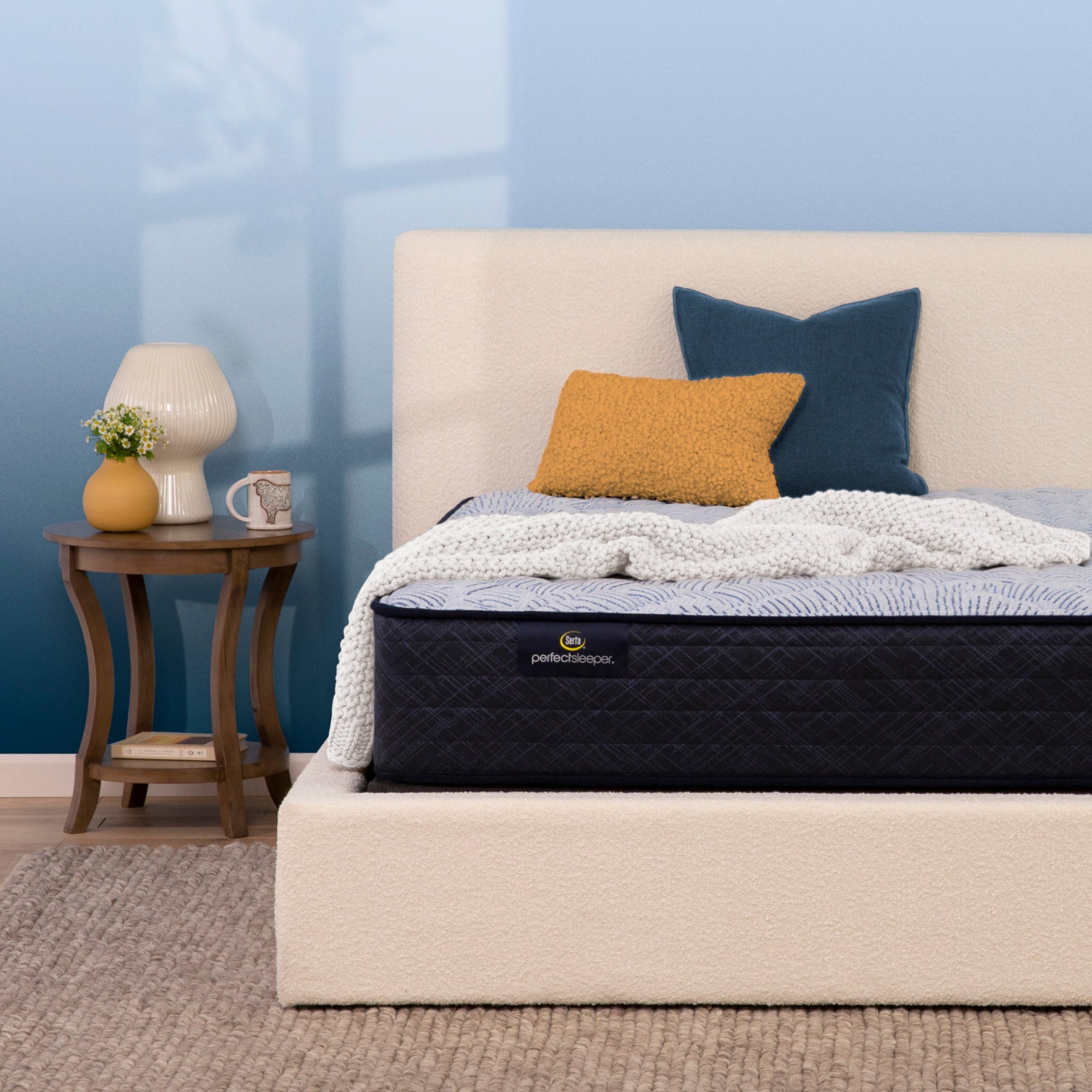 A Serta Perfect Sleeper firm mattress with a dark quilted, white textured bedding, and two pillows (one mustard yellow, one navy blue) in a styled bedroom setting with a blue wall, beige headboard, and a wooden side table.