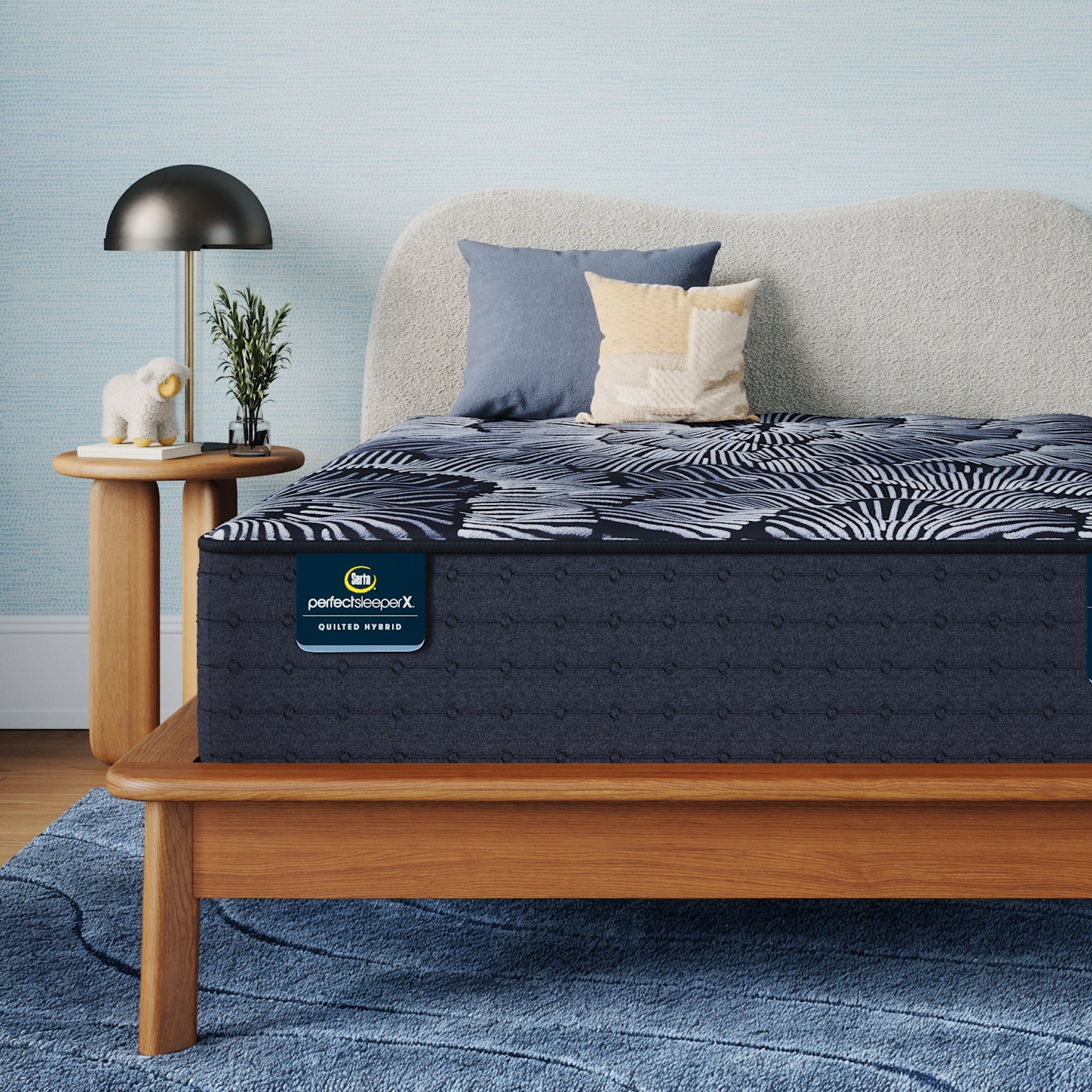 A Perfect Sleeper X Quilted Hybrid Excelled Plush mattress with a dark pattern and matching pillow shams styled in a bedroom setting, with a light wood bed frame, blue rug and side table