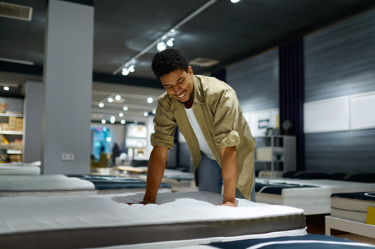 A man in a mattress store smiling at a mattress and pressing firmly with both hands onto the mattress.