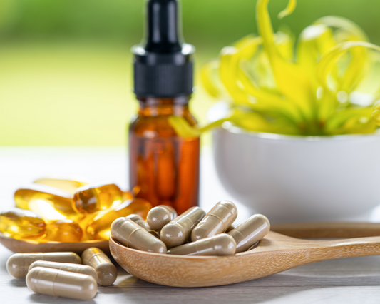 Daily vitamins in capsule format in a wooden spoon with a liquid vitamin dropper bottle and small plant in the background.