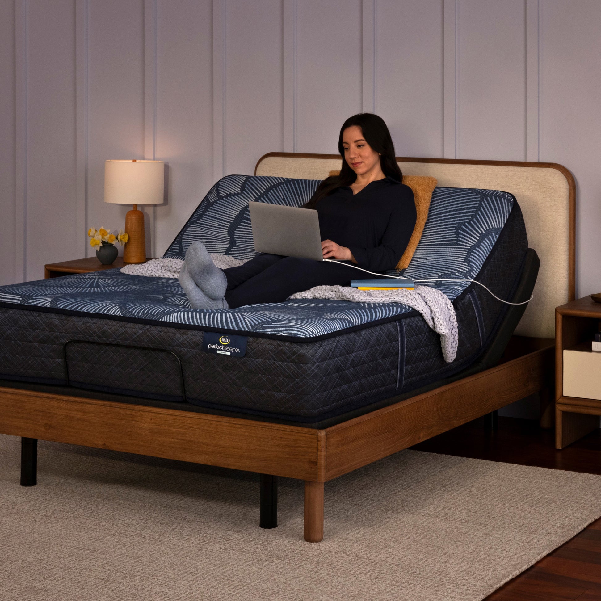 Woman siting on a perfect sleeper mattress slightly elevated at the top in the adjustable base, using her laptop that is plugged into the charging port on the side of the adjustable base.