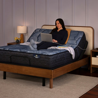 Woman siting on a perfect sleeper mattress slightly elevated at the top in the adjustable base, using her laptop that is plugged into the charging port on the side of the adjustable base.