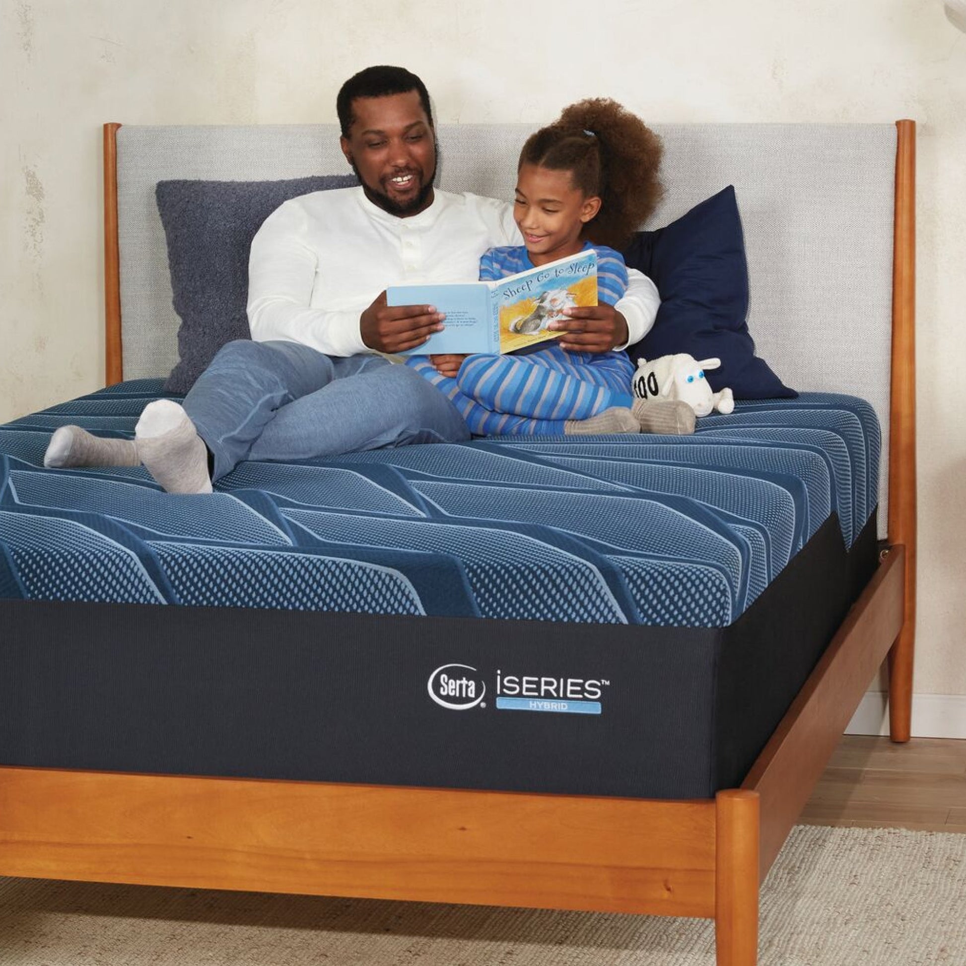 A man and little girl sitting on a Serta iSeries firm blue mattress, placed in a brown bed frame, reading a book while leaning on blue and grey pillows||feel: firm||level: enhanced